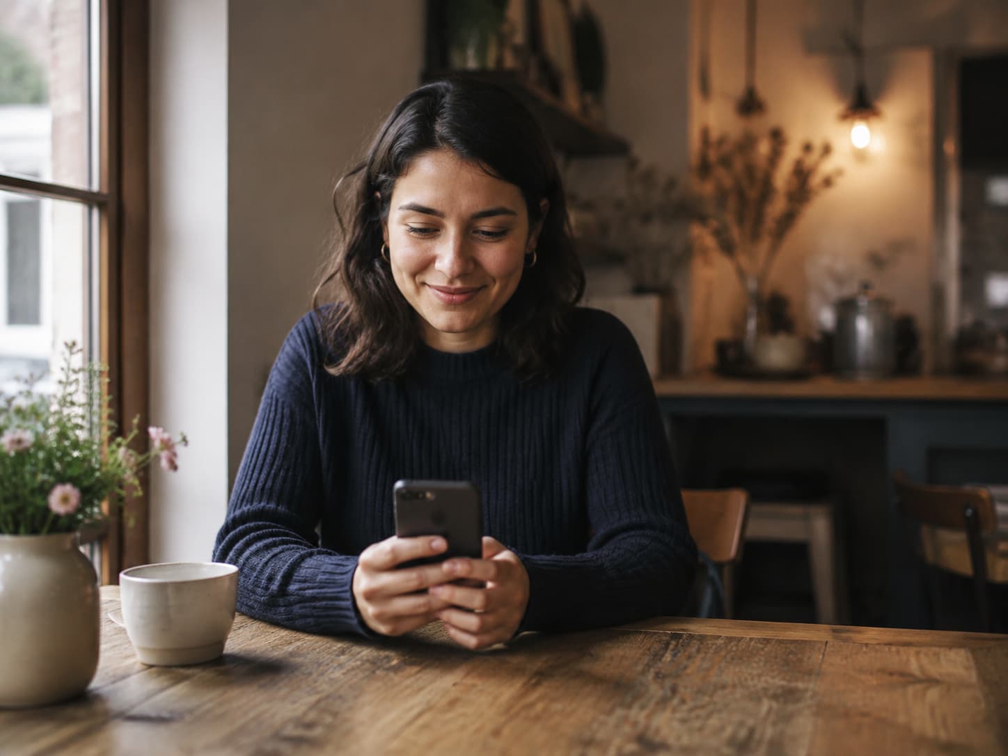 A woman smiling while checking a phone at a window table