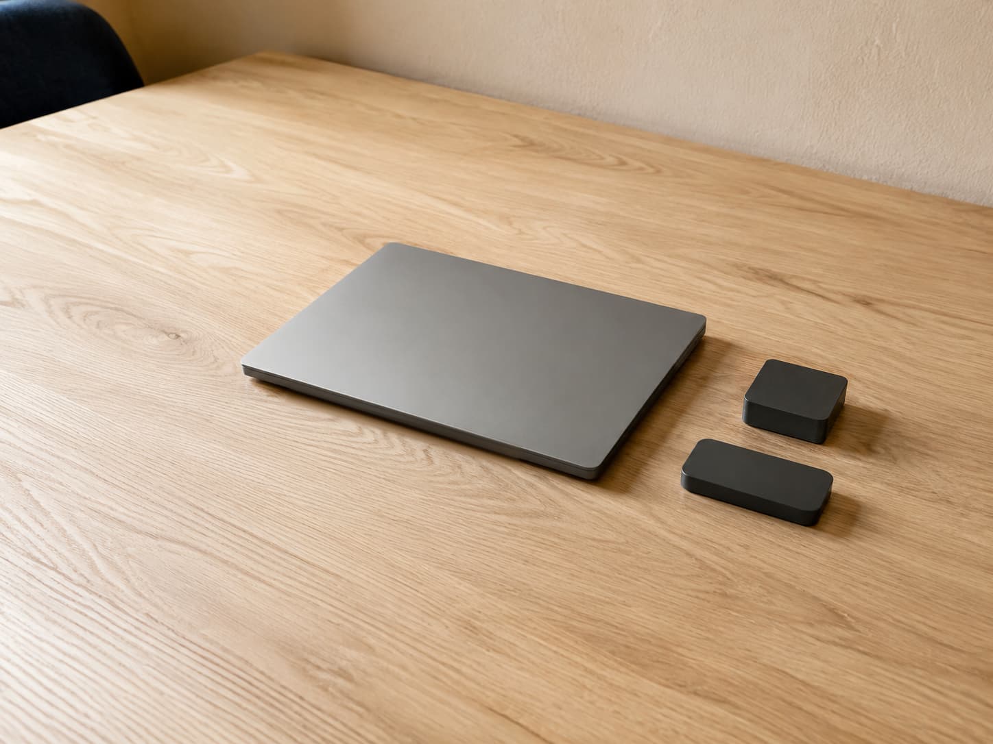 A closed silver laptop sits on a light wooden table beside a compact black charger and a slim portable storage drive.