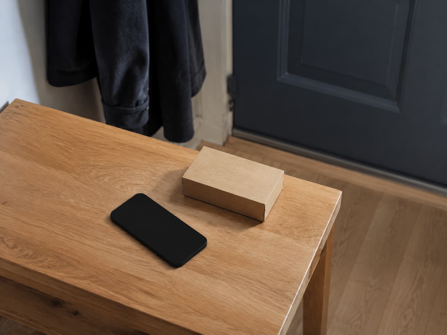 A black smartphone with its screen off and a small plain cardboard box sit on a light wooden table beside a dark front door and hanging coat.