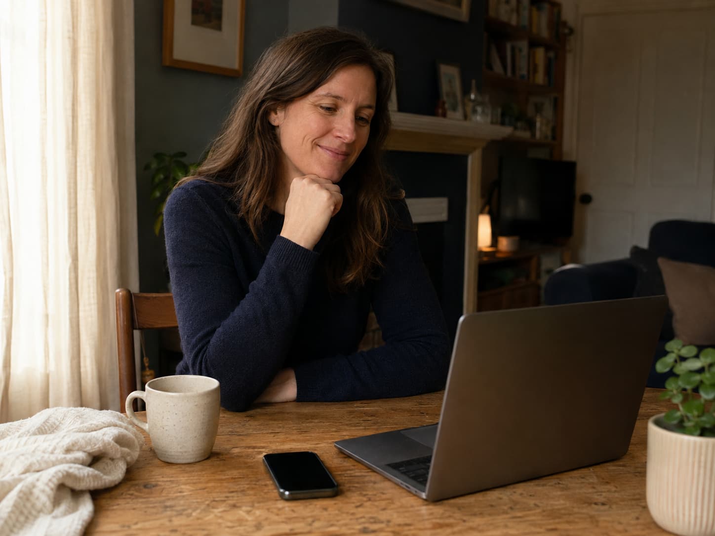 A woman looking at an open laptop beside a screen-off phone on a home table